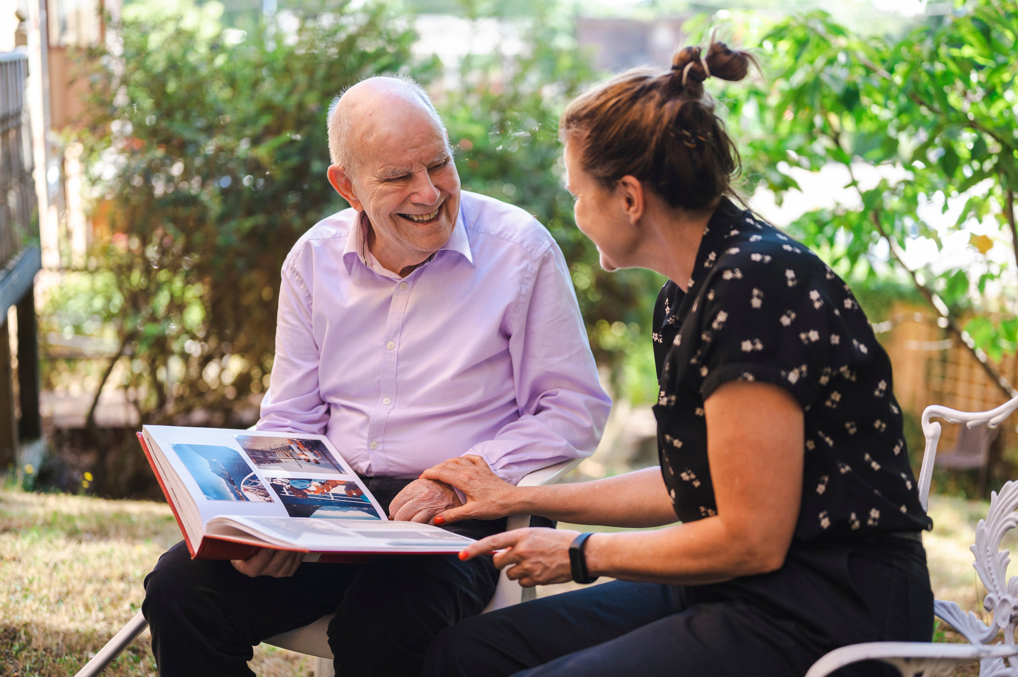 Smiling Older Man And Woman Sitting In Garden Looking Through Photo Album