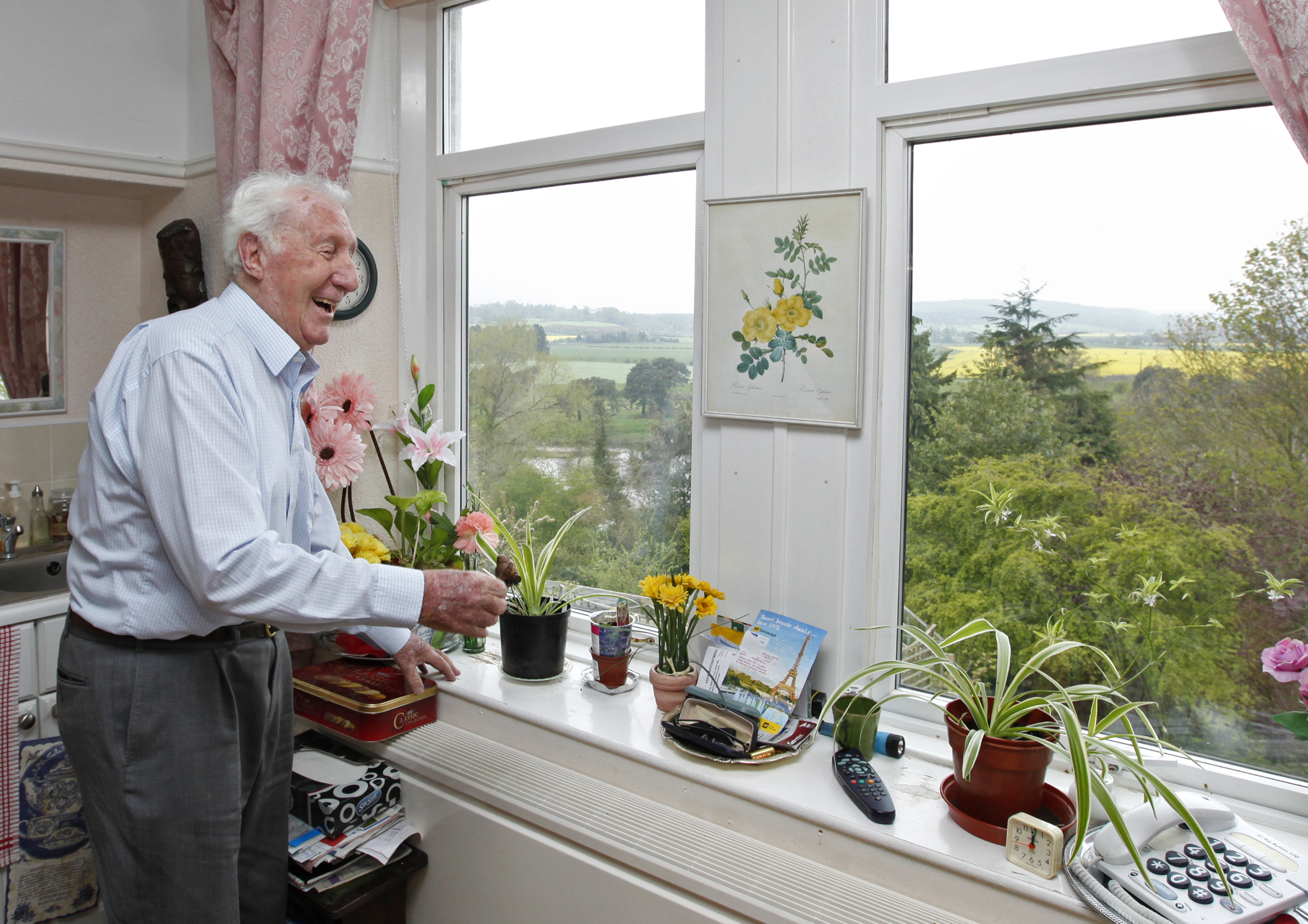 A gentleman is stood in the window looking at plants on the sill