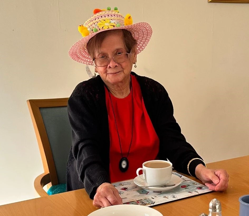 An older lady sitting down with a cup of tea and an easter bonnet on her head