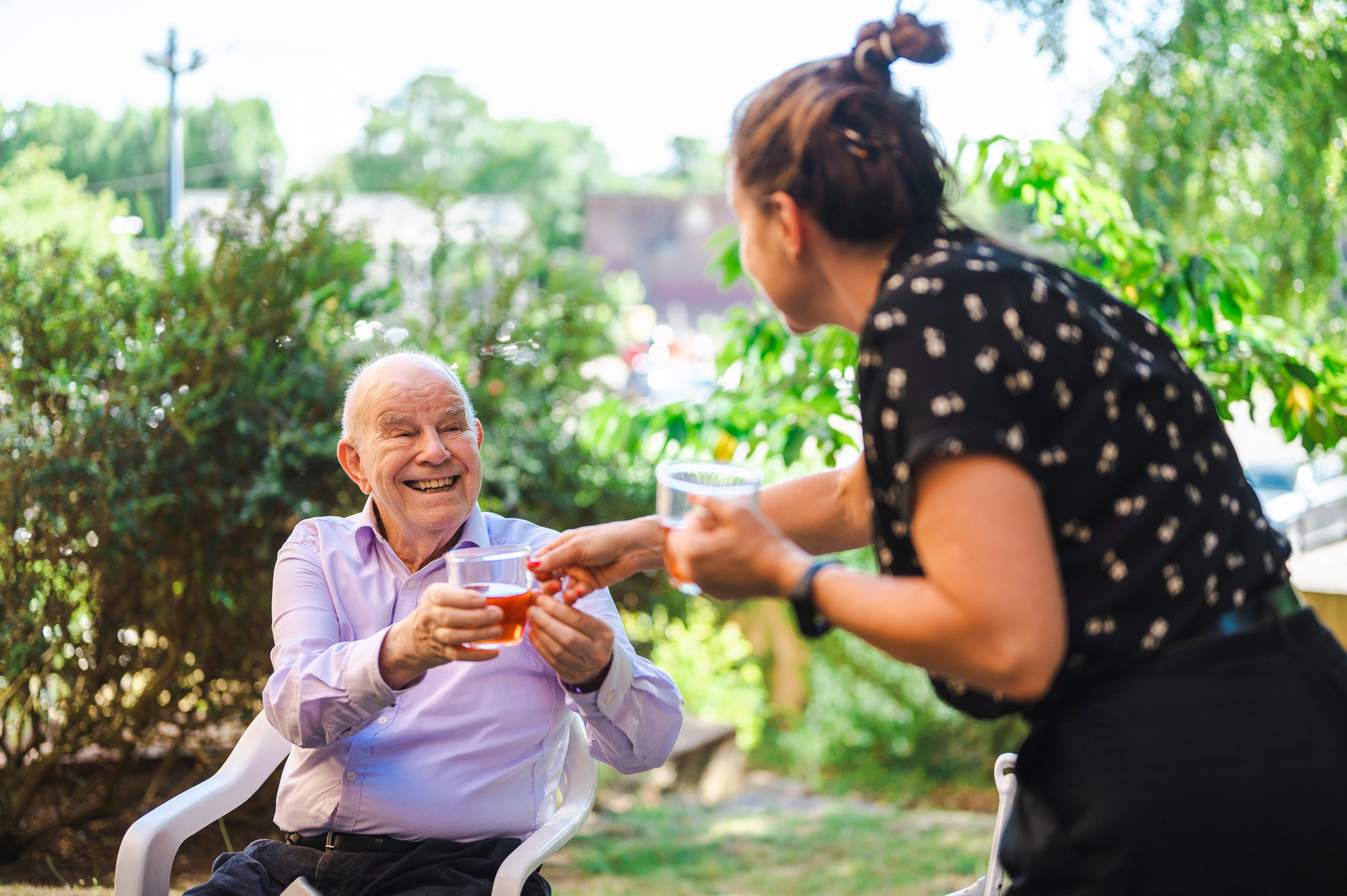 Older Man Sitting In Garden Smiling And Being Handed A Drink By A Woman