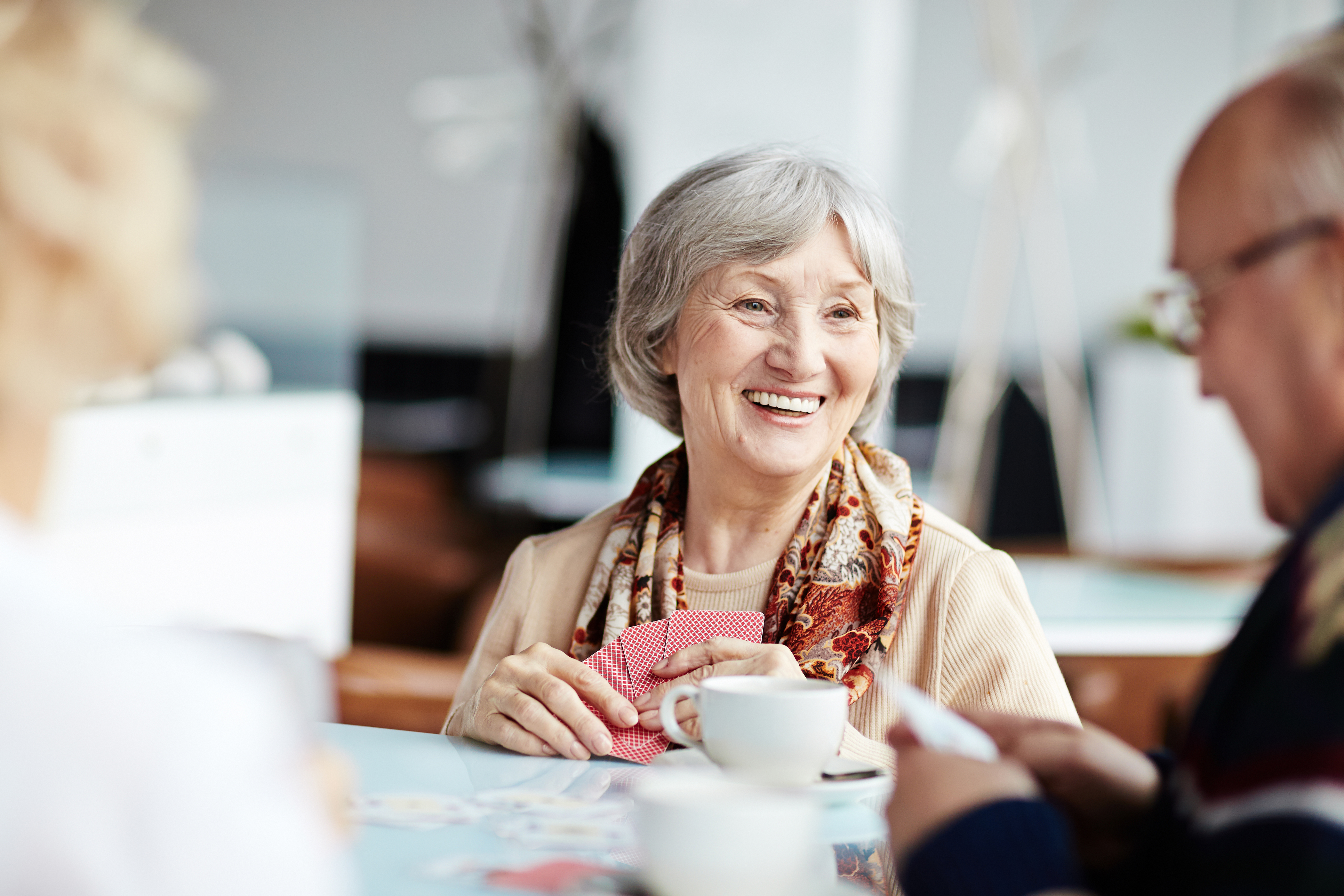 Smiling older woman enjoying playing cards around a table with others with a cup of tea