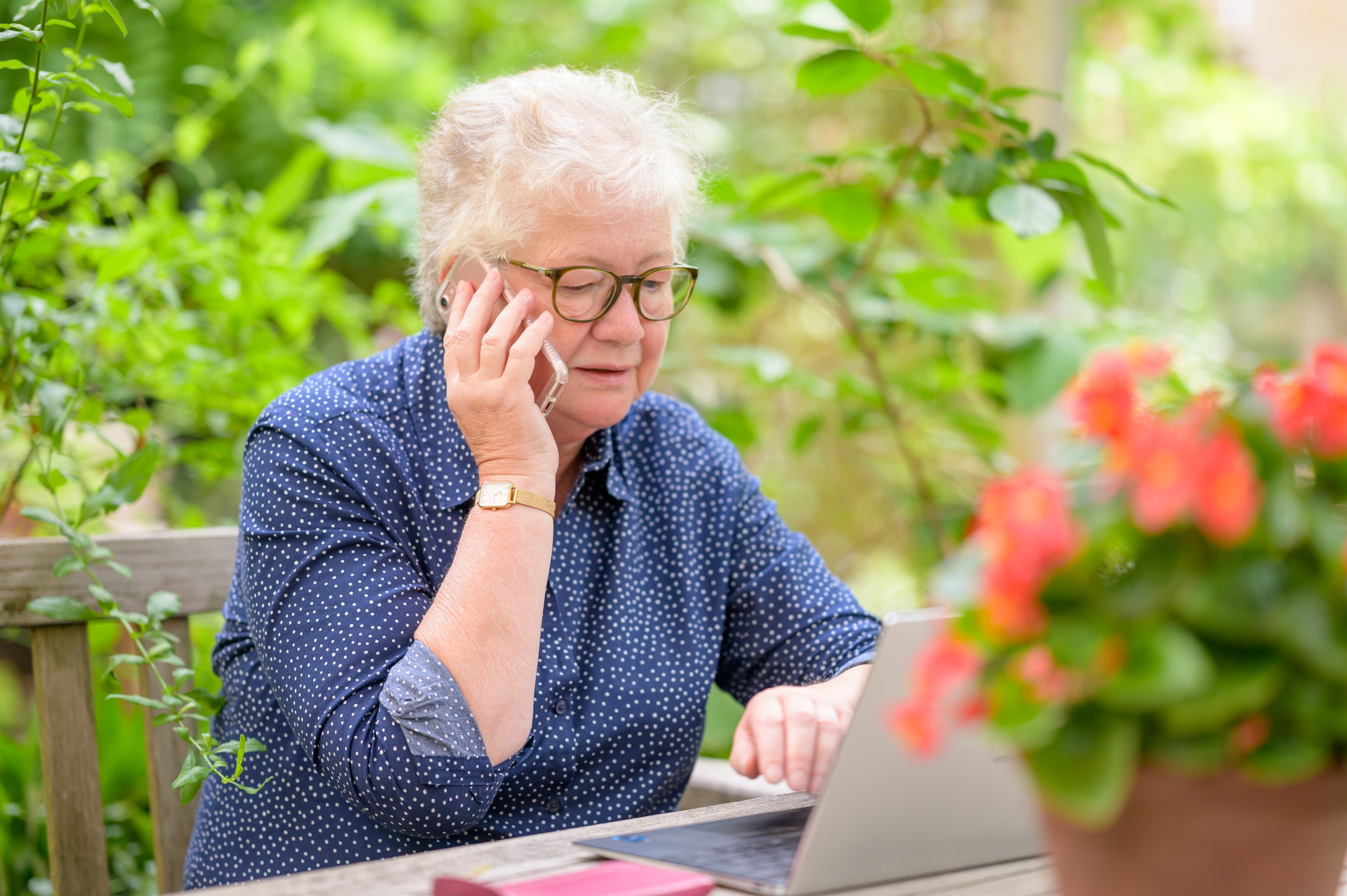 Older Woman In Garden Working On Her Laptop And On The Phone