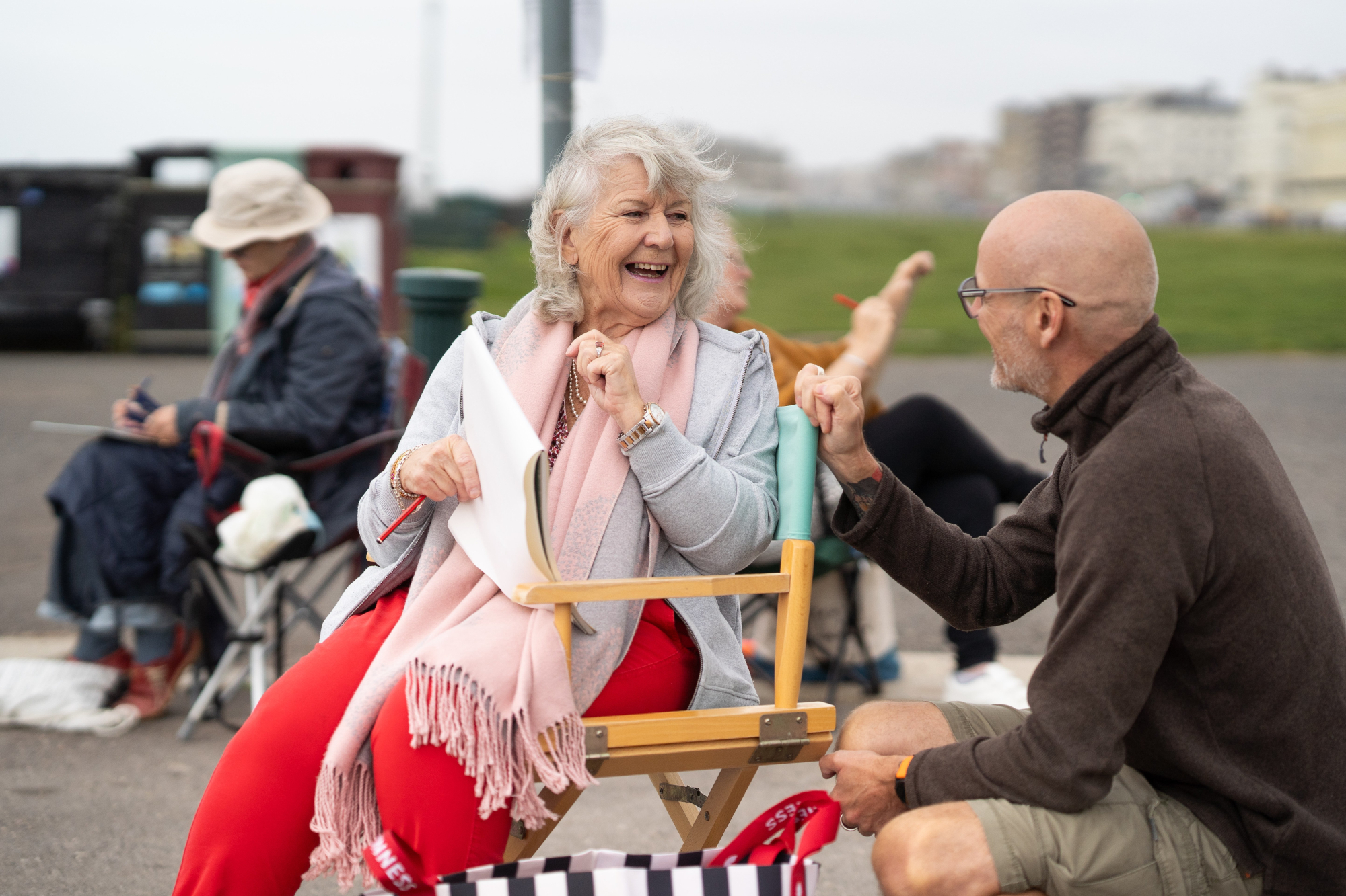 Older Woman Outside Sitting In Chair Smiling Chatting To Another Man