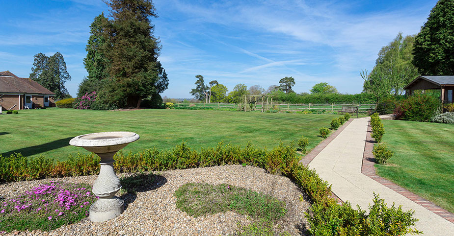 Expansive gardens at Westall House featuring a stone bird bath, a winding pathway, a large lawn, and mature trees under a blue sky with clouds