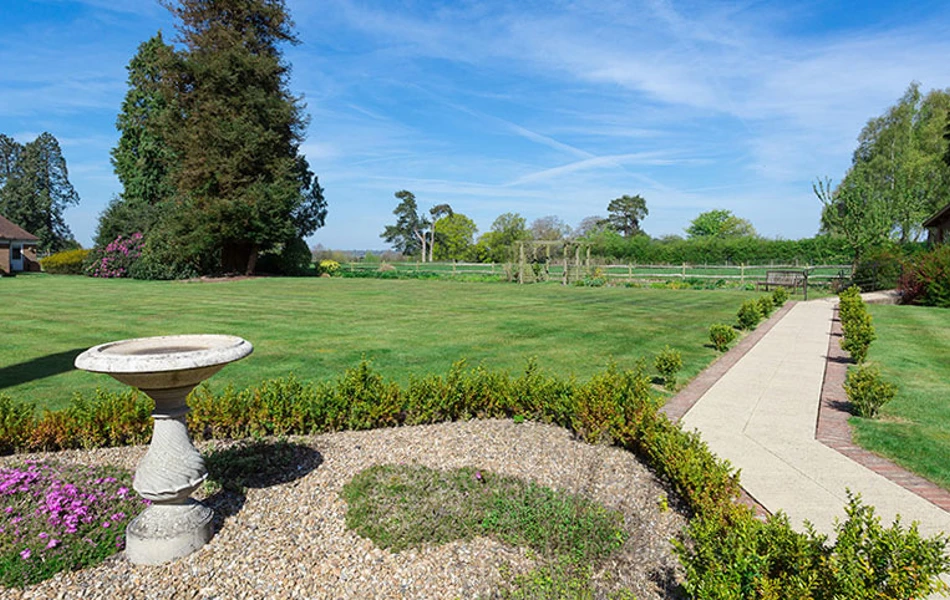 Expansive gardens at Westall House featuring a stone bird bath, a winding pathway, a large lawn, and mature trees under a blue sky with clouds