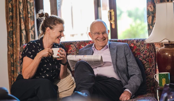 Woman And Older Man Smiling On The Sofa With A Cup Of Tea And A Newspaper