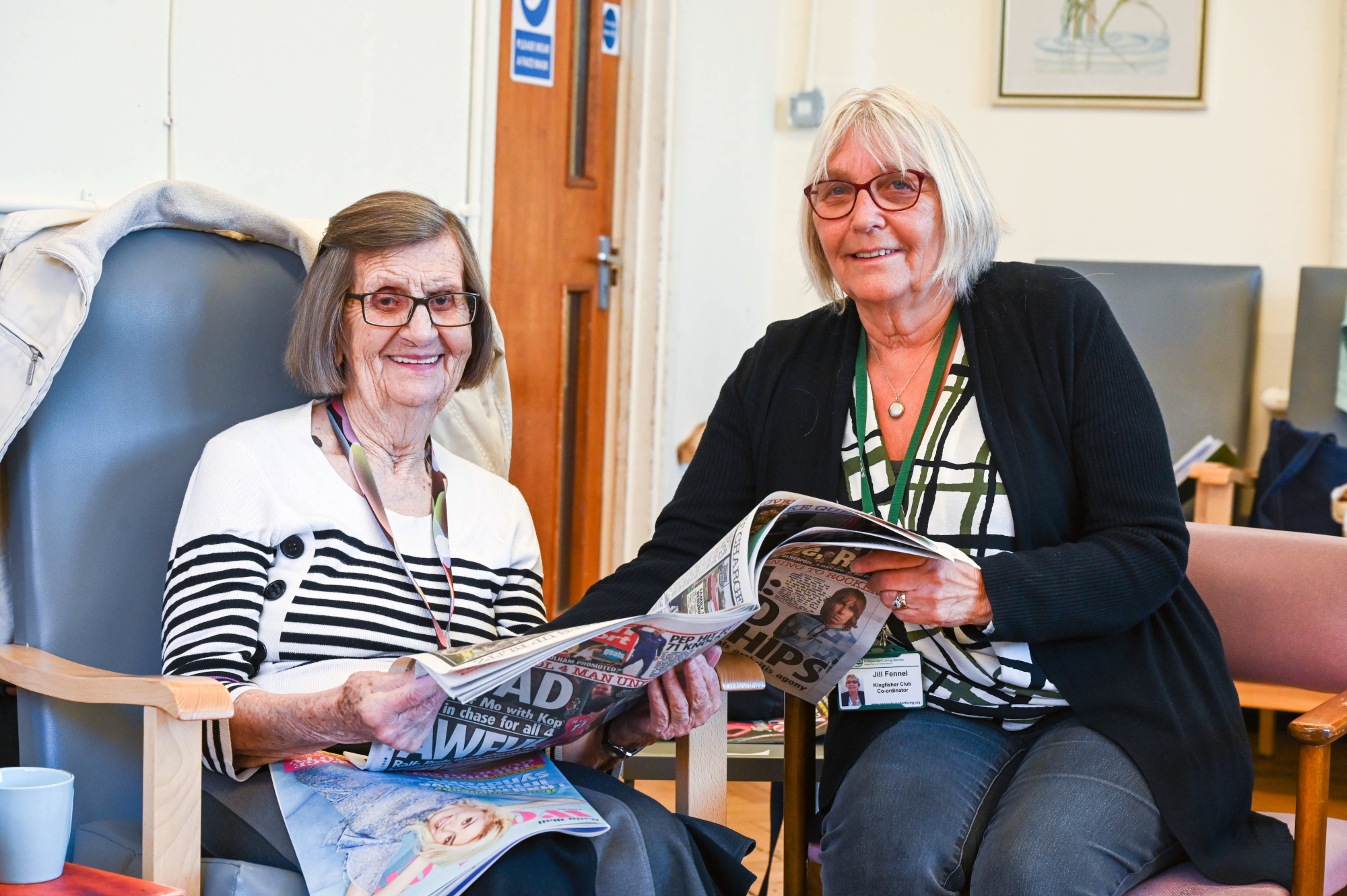 Older Woman And Staff Member Looking Through The Newspaper Together